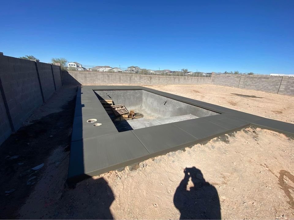 A rectangular concrete pool under construction in an open, sandy area showcases the expertise of Mesa Master Concrete. The empty basin, dotted with construction materials, stands under a clear blue sky with a partially built gray wall surrounding the scene.