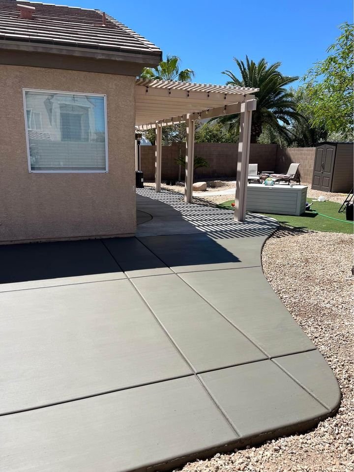 A backyard scene in Mesa AZ features a freshly paved cement path curving around a single-story house. A wooden pergola provides shade over outdoor furniture, surrounded by gravel, a grassy area, and palm trees. Expertise from local concrete contractors is evident. A fence and storage shed are in the background.