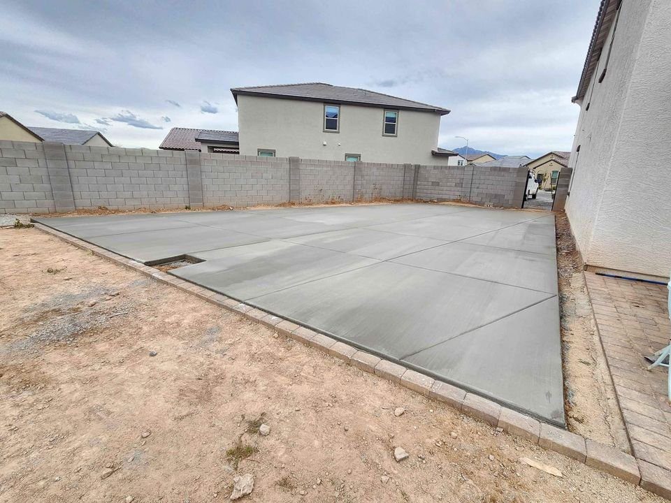 In Apache Junction, a freshly poured concrete slab graces the backyard, encircled by a low brick wall. Two houses stand under a cloudy sky, and dirt surrounds the slab. For comprehensive concrete services and a free quote, this transformation could be yours.