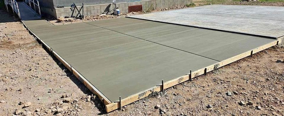 A freshly poured concrete slab is framed with wooden planks in an outdoor setting in Mesa AZ. Surrounded by rocky ground, the concrete appears smooth and neatly sectioned into squares, reflecting the meticulous work of a skilled foundation contractor.