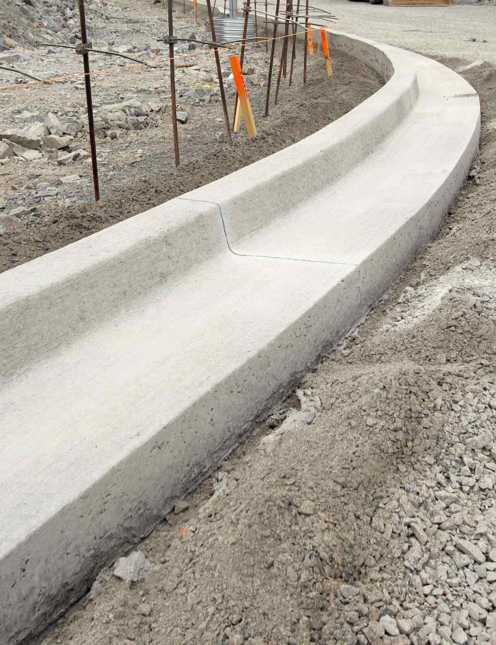 A new concrete curb under construction, crafted by expert concrete contractors, is surrounded by gravel and dirt. Scaffolding and safety markers are visible in the background, ensuring a safe environment for this Mesa AZ project.