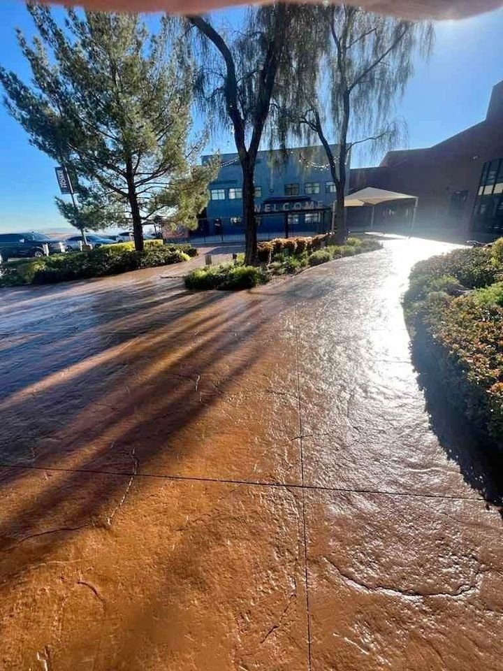 A sunlit walkway with textured paving, part of a concrete construction typical in Mesa AZ, leads towards a building partially shaded by trees. The morning sun casts long shadows, creating a warm and inviting atmosphere. Bushes line the path, and a sign on the building says Welcome.