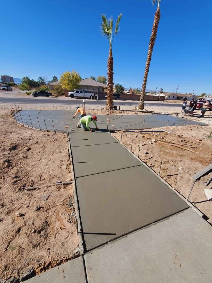 In sunny Mesa, contractors skillfully smooth wet concrete on a newly laid sidewalk. The path leads to a circular concrete pad, surrounded by palm trees and parked cars in the background.