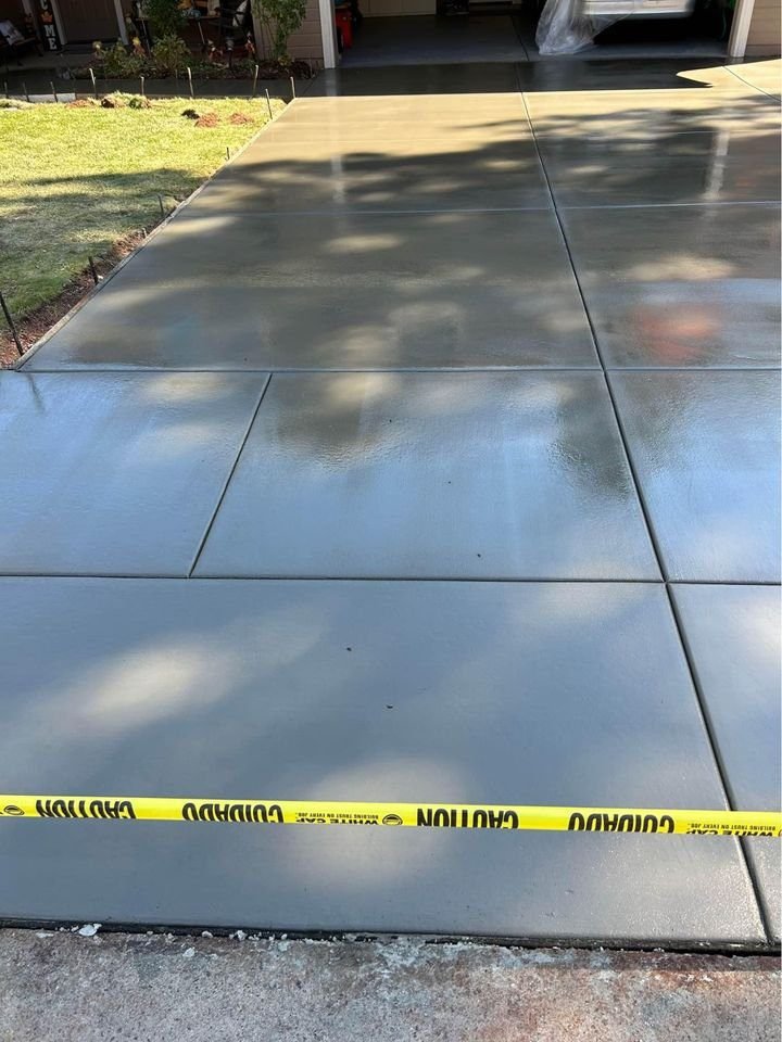 Freshly poured concrete driveway glistens with a smooth, shiny surface. Yellow caution tape stretches across the area to ensure safety during the concrete driveway installation. In the background, grass and a garage hint at another successful project by Mesa Master Concrete.