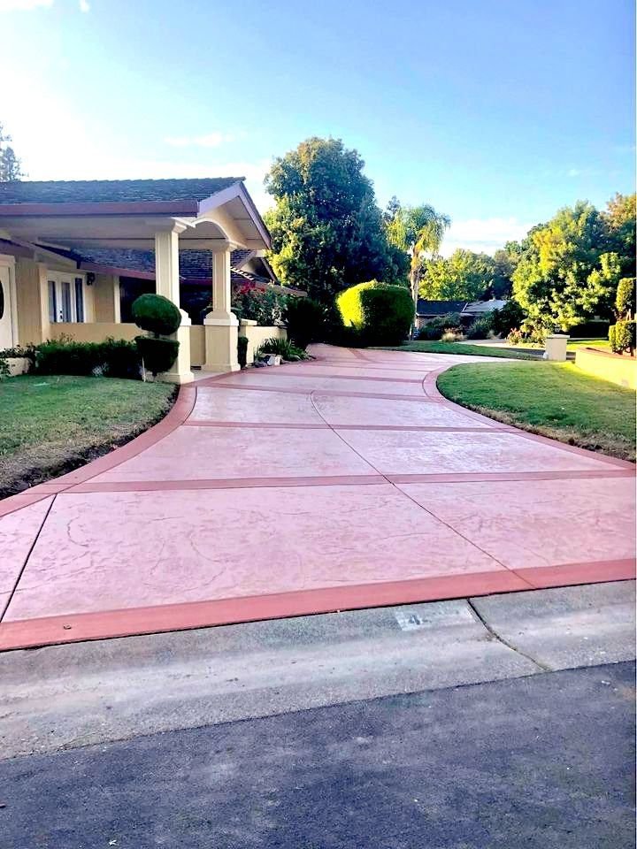 A house with a long, curved concrete driveway lined with grass and shrubs welcomes you. The Mesa, AZ sun casts a warm glow on its reddish hue, leading to the entrance. The front yard is well-maintained, featuring trees and bushes under a clear blue sky.