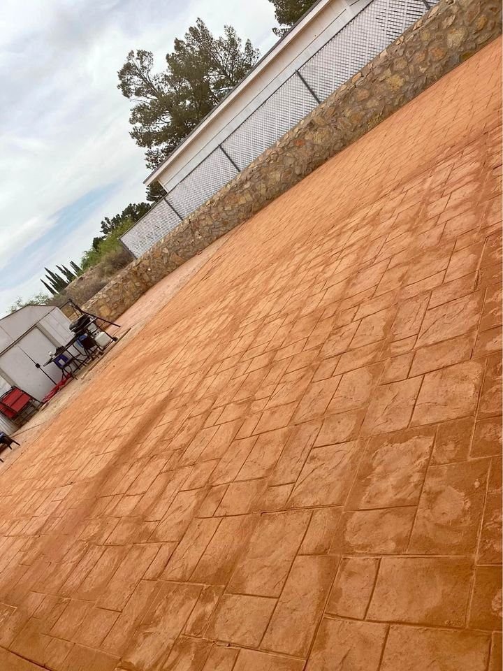 A large, empty patio with a textured orange-brown surface is shown. In the background, there's a stone wall and metal fence, with trees and a cloudy sky typical of Mesa. Some chairs and equipment from a recent Mesa Master Concrete driveway installation are visible on the left side.