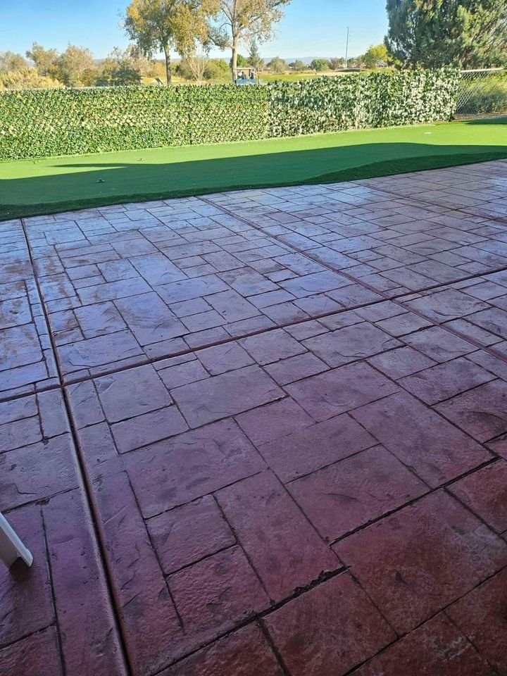 A wet red-tiled patio with a green artificial turf lawn in the background, crafted with precision by Mesa Master Concrete. The scene is bordered by a tall hedge, and several trees are visible in the distance under a clear blue sky.