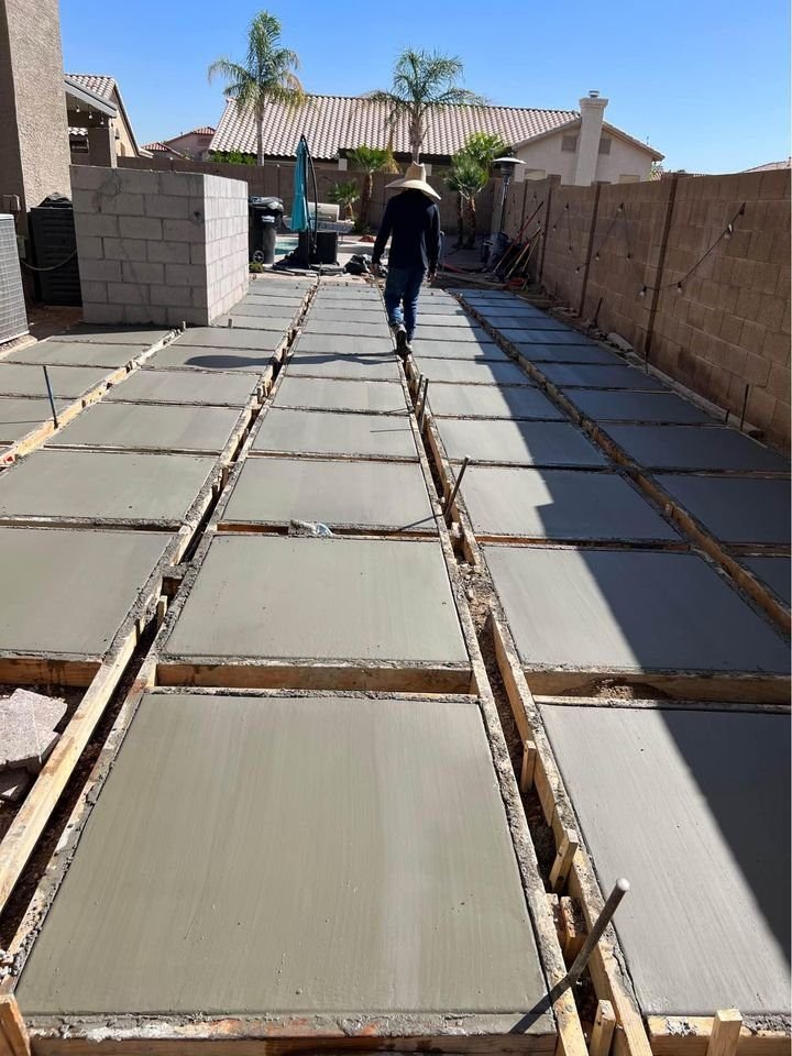 A person walks between large, freshly laid concrete slabs in a Mesa AZ backyard under a clear blue sky. The slabs are framed with wooden planks and lined up in rows. Palm trees and neighboring houses are visible in the background, showcasing the skill of a local concrete contractor.