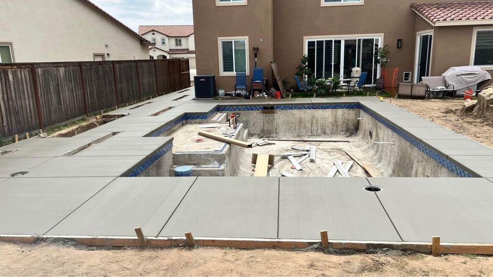 A backyard pool under construction showcases the expertise of concrete contractors in Tempe, AZ, with freshly poured concrete surrounding it. The pool interior is unfinished with construction materials. Nearby, patio furniture and a grill sit by the two-story house featuring tan exterior walls.