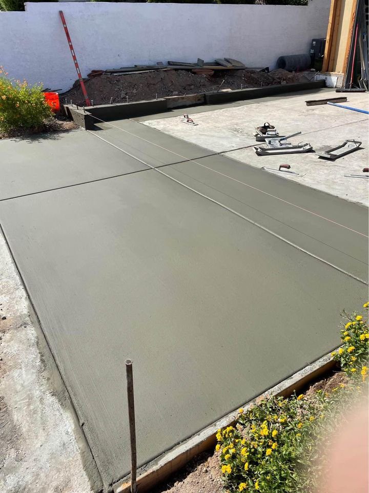 In a Tempe, AZ backyard, freshly poured concrete solutions create a smooth, even slab bordered by small yellow flowers. Construction tools lie scattered nearby, while a red construction marker stands against the white wall in the background—a testament to skilled concrete contractors at work.