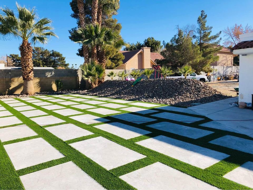 A landscaped backyard in Tempe, AZ features a geometric pattern of concrete slabs with lush green grass in between. Palm trees surround the area, with a pile of rocks near a house boasting a red tile roof. The clear, sunny sky completes this serene scene crafted by expert concrete contractors.