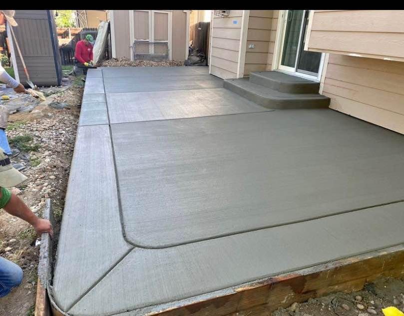 Workers are smoothing freshly poured concrete on a backyard patio next to a house, showcasing expertise common in Scottsdale projects. The surface gleams with a smooth finish, and the edges are well-defined. Steps lead to a door, while construction equipment from Arizona-licensed concrete contractors is nearby.