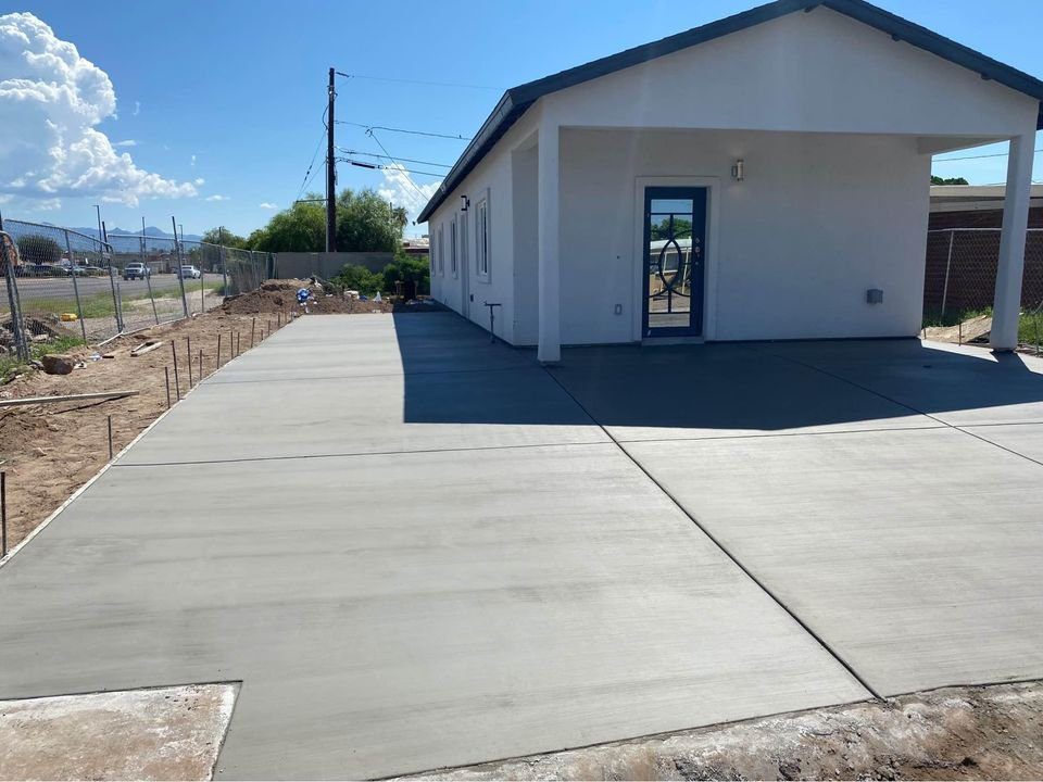 A freshly paved concrete driveway, courtesy of Mesa Master Concrete, leads to a white house with a small overhang and blue-trimmed windows. The clear sky has scattered clouds above the fenced area with visible utility poles and a dirt pathway beside it in charming Chandler, AZ.