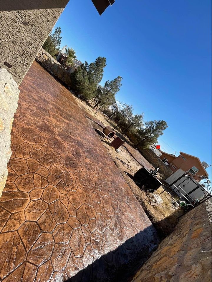 A backyard with a textured concrete patio that resembles stone. Trees and a brick house with a satellite dish are in the background under the clear Tempe, AZ sky. Gardening tools and a wheelbarrow are also visible near this inviting patio—a testament to quality concrete solutions.