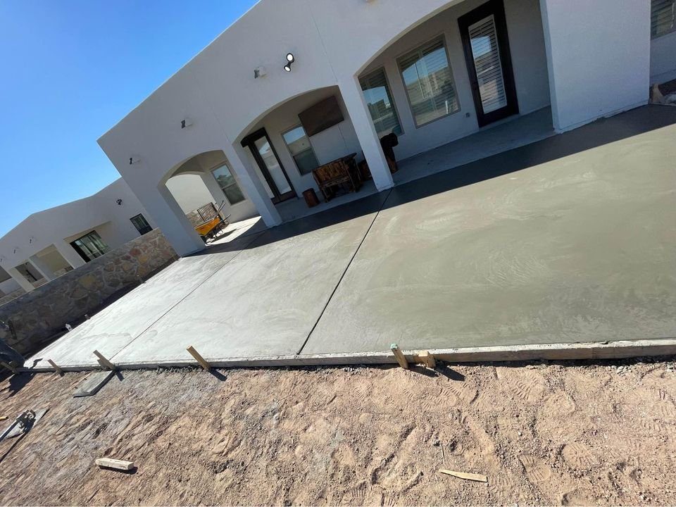 A newly poured concrete patio in front of a white house with arched doorways and windows showcases the expertise of leading concrete contractors in Tempe, AZ. The patio is surrounded by a dirt area with wooden stakes, all under a clear, blue sky.